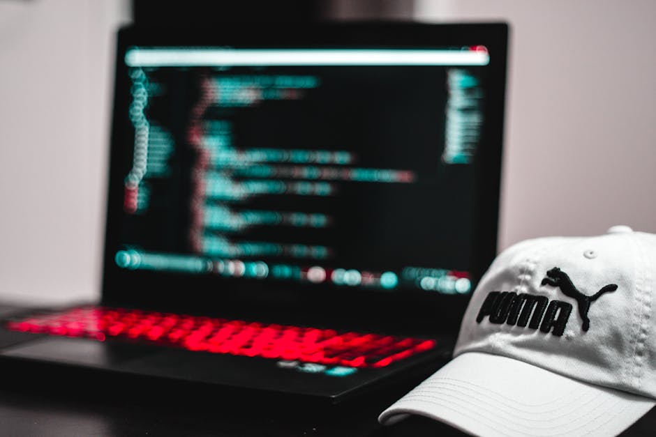 Close-up of a person's hands operating a laptop on a dark surface, displaying a code editor with lines of programming code in various colours, inside a home or office environment with soft lighting. T
