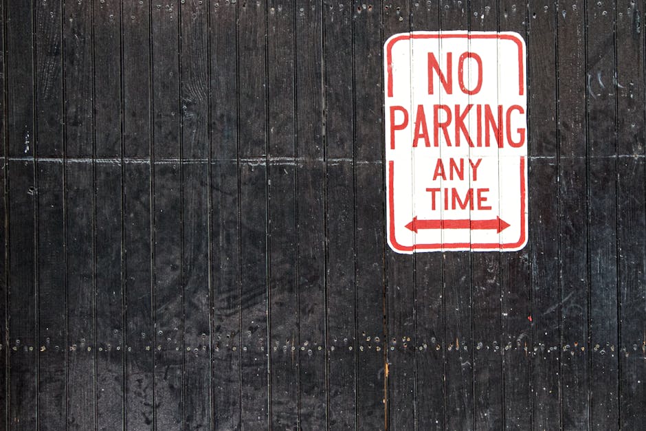 Close-up view of a black wooden wall with vertical planks, featuring a white and red 'No Parking Any Time' sign attached to the surface. The sign has red text and borders, with two red arrows pointing