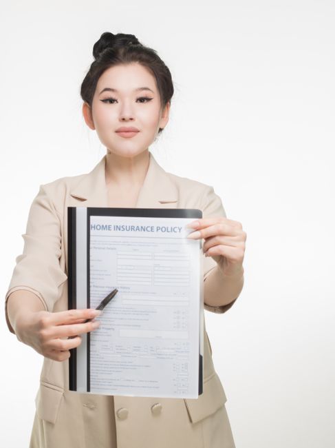 A woman with dark hair styled into a neat updo, dressed in a beige blazer, holds a clipboard displaying a paper titled 'Home Insurance Policy' and a pen. She is standing against a plain white backgrou