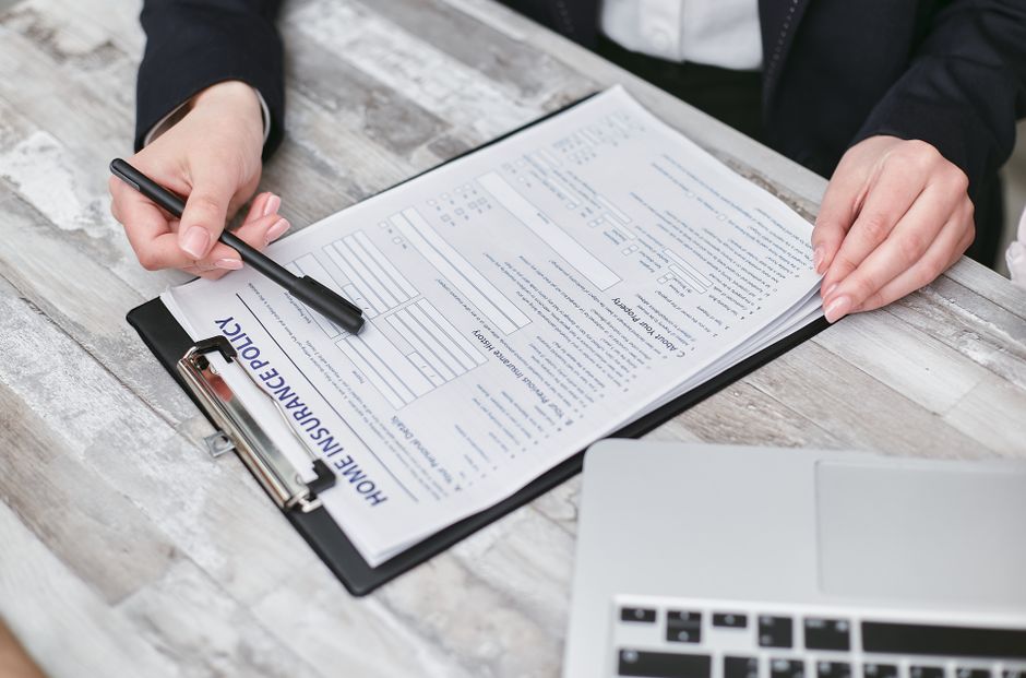 A woman with dark hair styled into a neat updo, dressed in a beige blazer, holds a clipboard displaying a paper titled 'Home Insurance Policy' and a pen. She is standing against a plain white backgrou