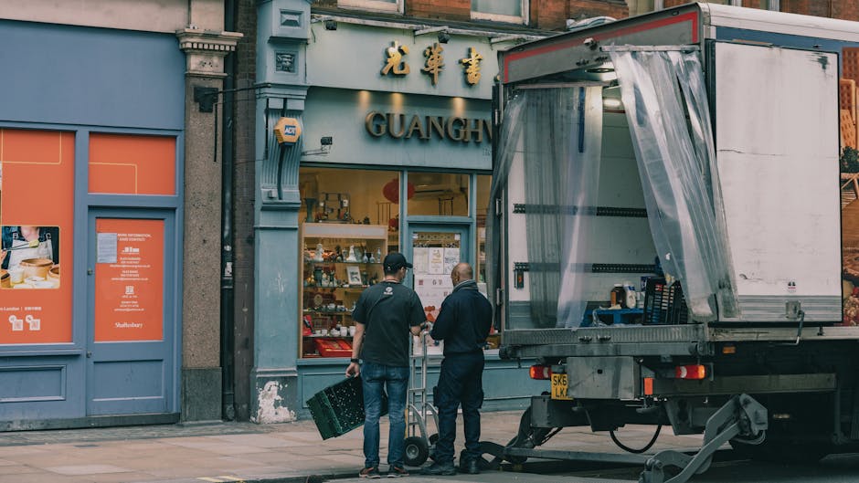 Two men are standing outside a shop with a sign displaying Chinese characters and the name 'GUAGE' on a blue-grey facade, preparing for a home relocation or furniture transport. One man, dressed in da